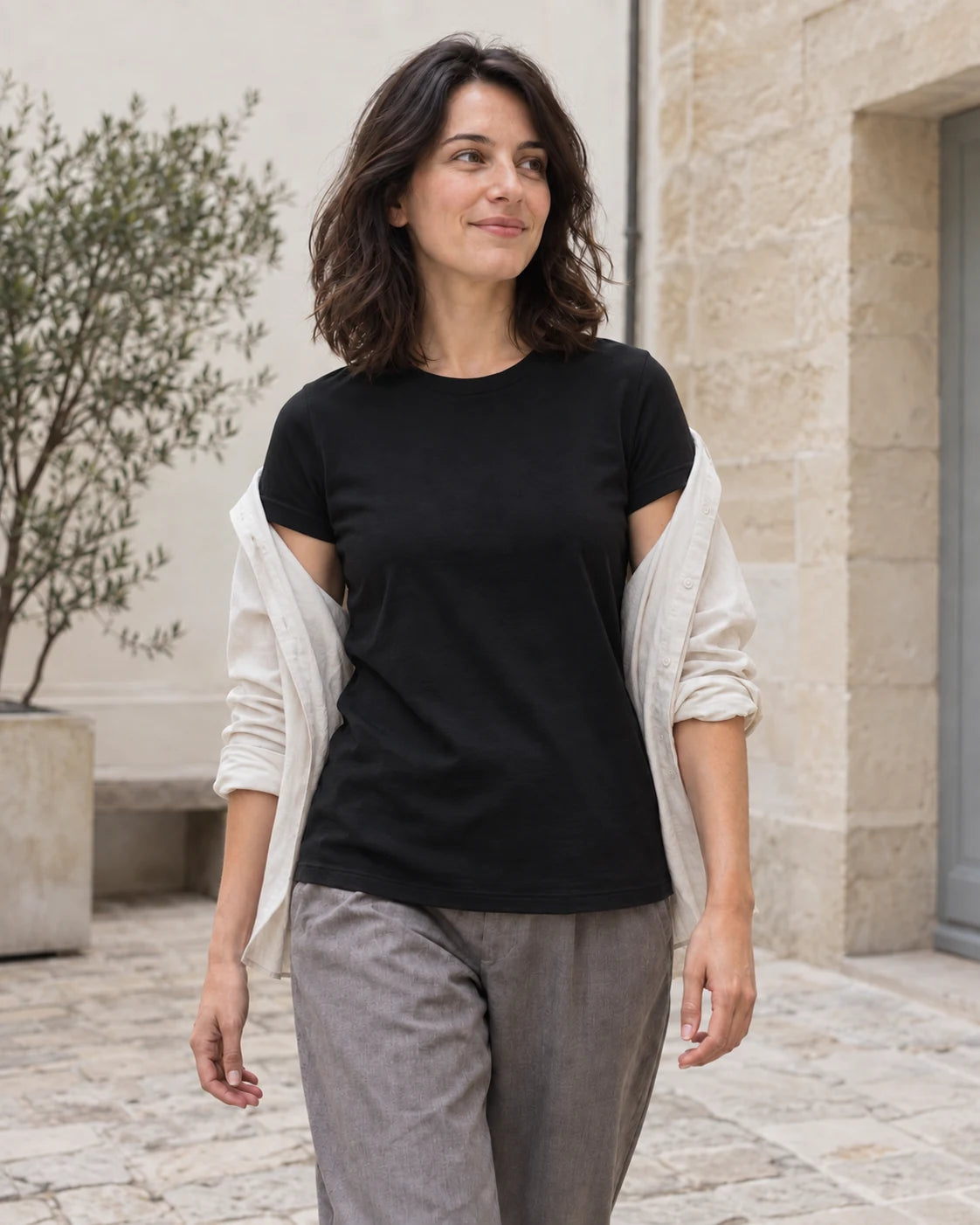 Woman wearing a black crew-neck tee with an open pale overshirt in a stone courtyard.