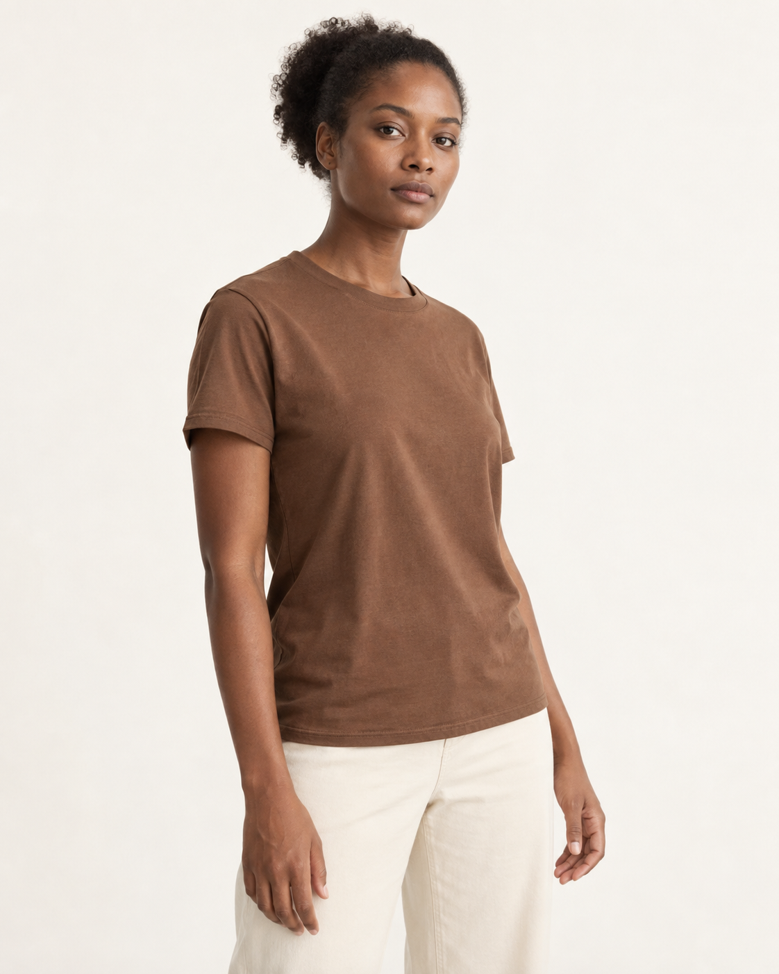 Woman in a brown crew-neck tee and ivory trousers, standing at a slight angle with a neutral expression in a studio.